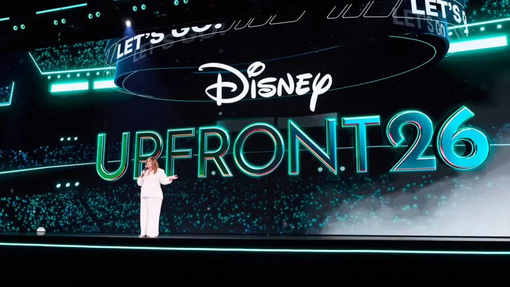 Presenter in a white suit speaks on a brightly lit stage with a large teal 'UP FRONT 26' backdrop and the Disney logo behind her, at a Disney Up Front event.