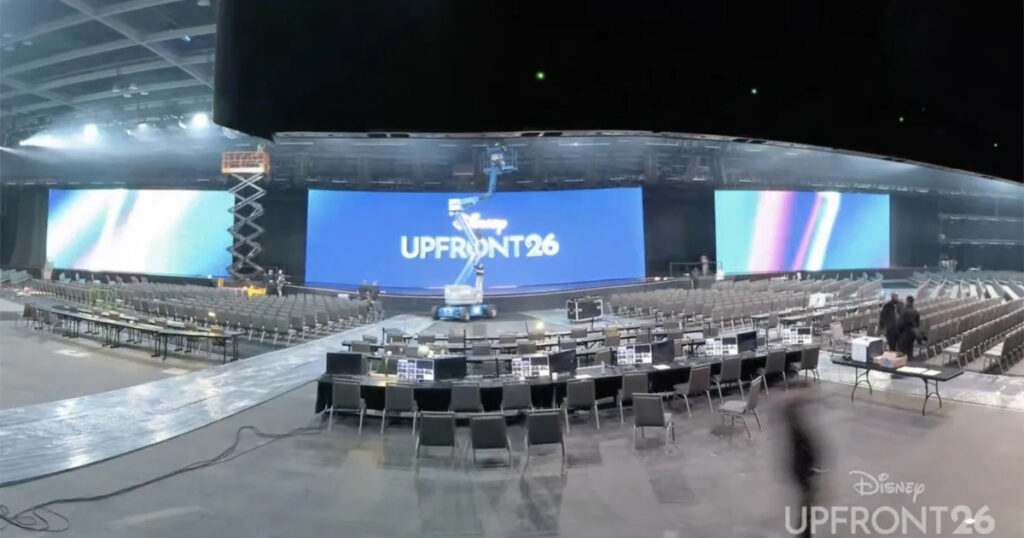 Wide shot of a large convention hall with a blue UPFRONT26 stage screen, rows of chairs, and crew preparing the setup.