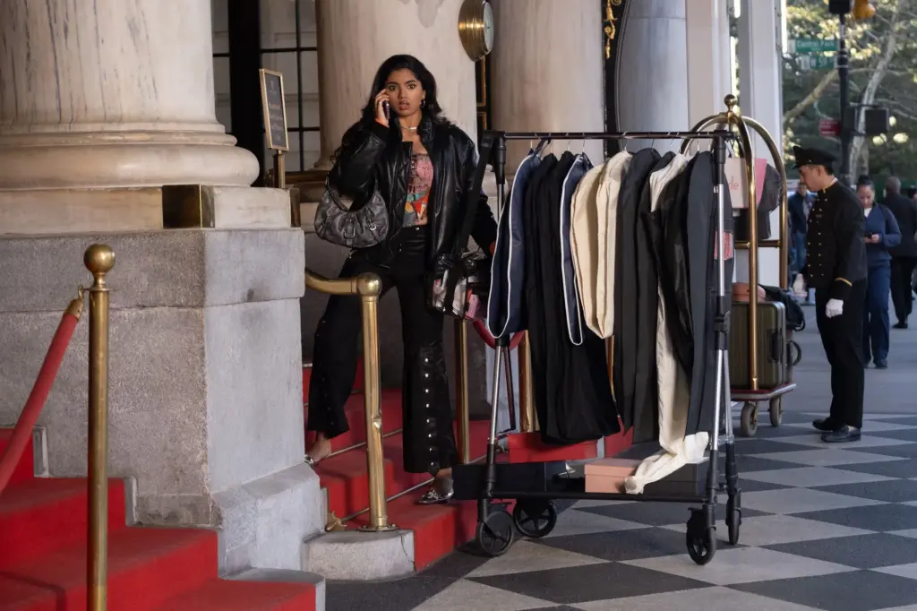 Woman on a red carpet outside a grand entrance, talking on her phone beside a rack of coats and a luggage cart nearby.